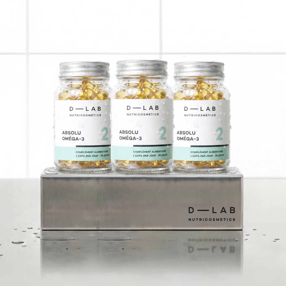 Absolu Oméga-3 D-LAB Nutricosmetics, cure 3 mois : 3 flacons de gélules alignés sur un socle inox brossé, fond carrelage blanc