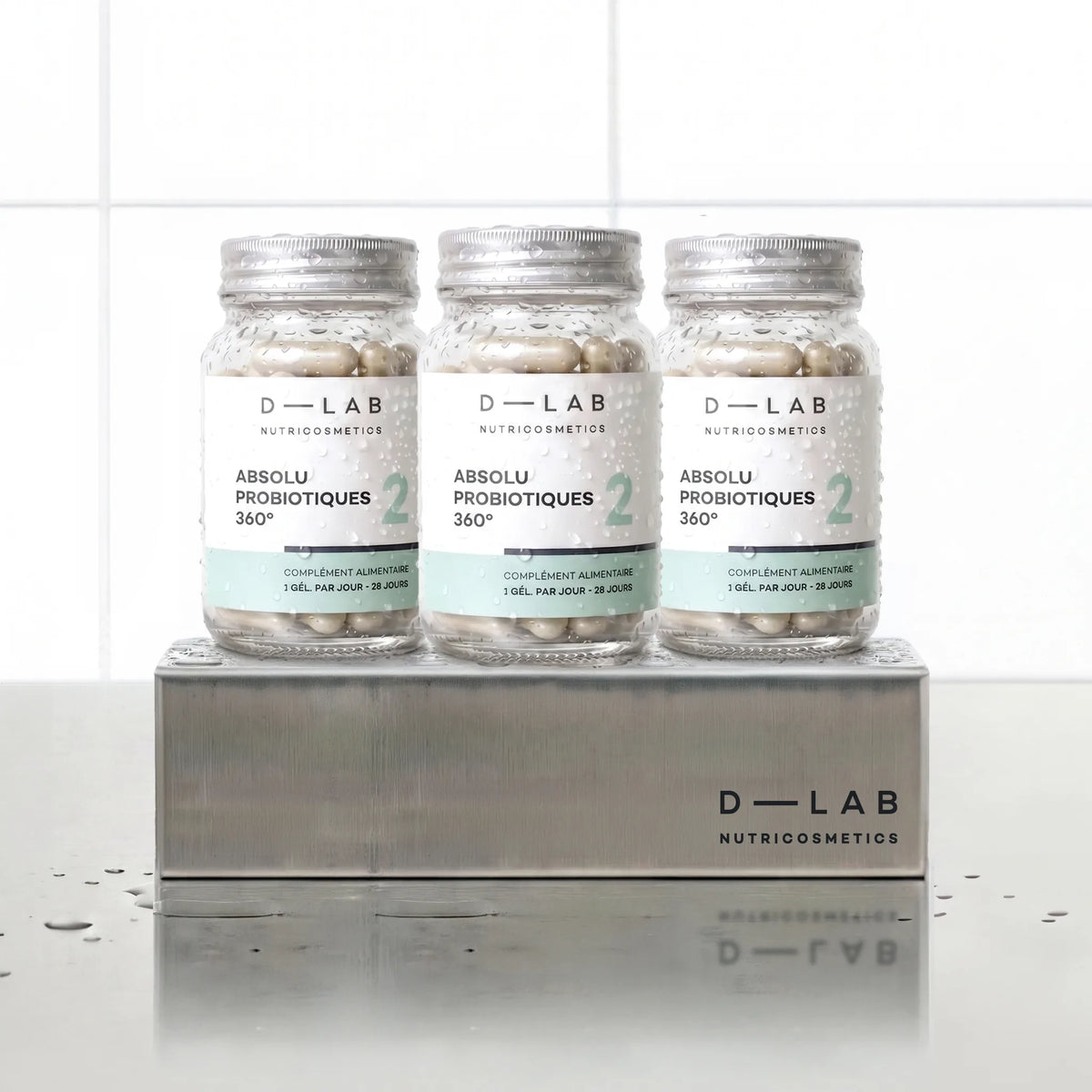 Absolu Probiotiques 360° D-LAB Nutricosmetics, cure 3 mois : 3 flacons de gélules alignés sur un socle inox brossé, fond carrelage blanc