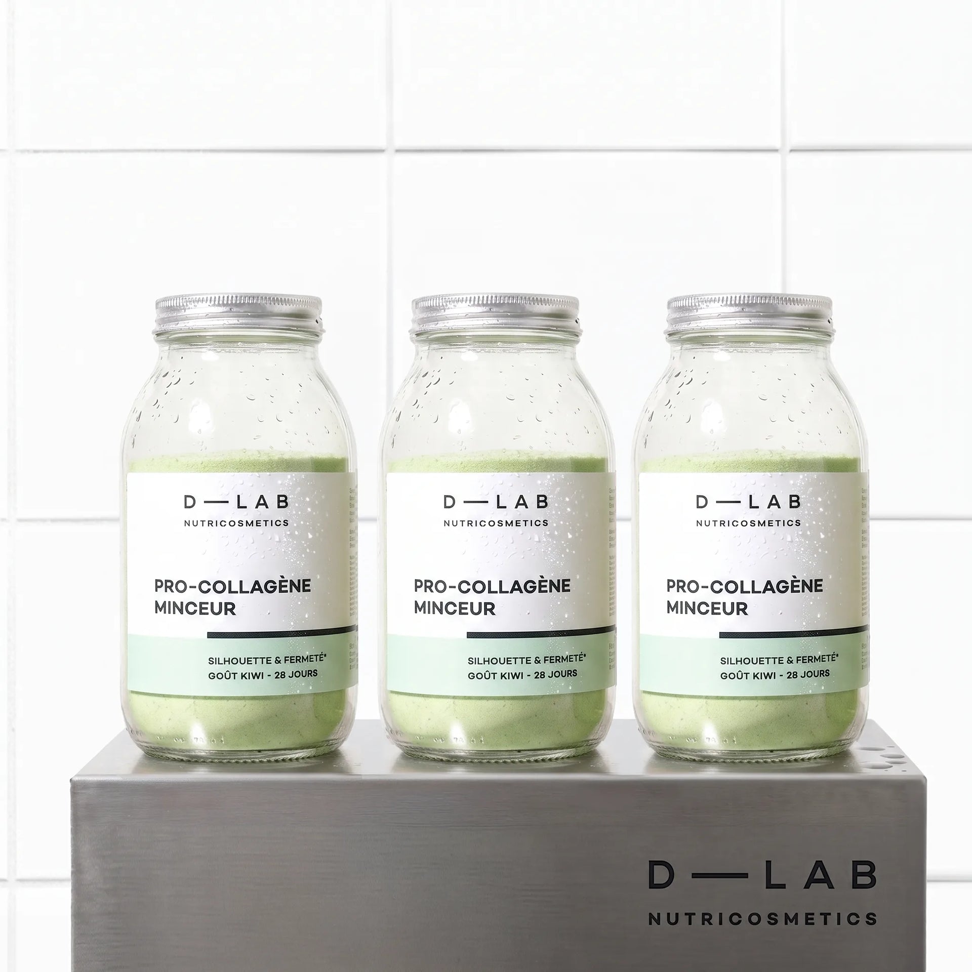 Cure 3 mois Pro-Collagène Minceur goût kiwi D-LAB, trois flacons en verre de poudre verte sur socle en métal brossé, fond carrelage blanc