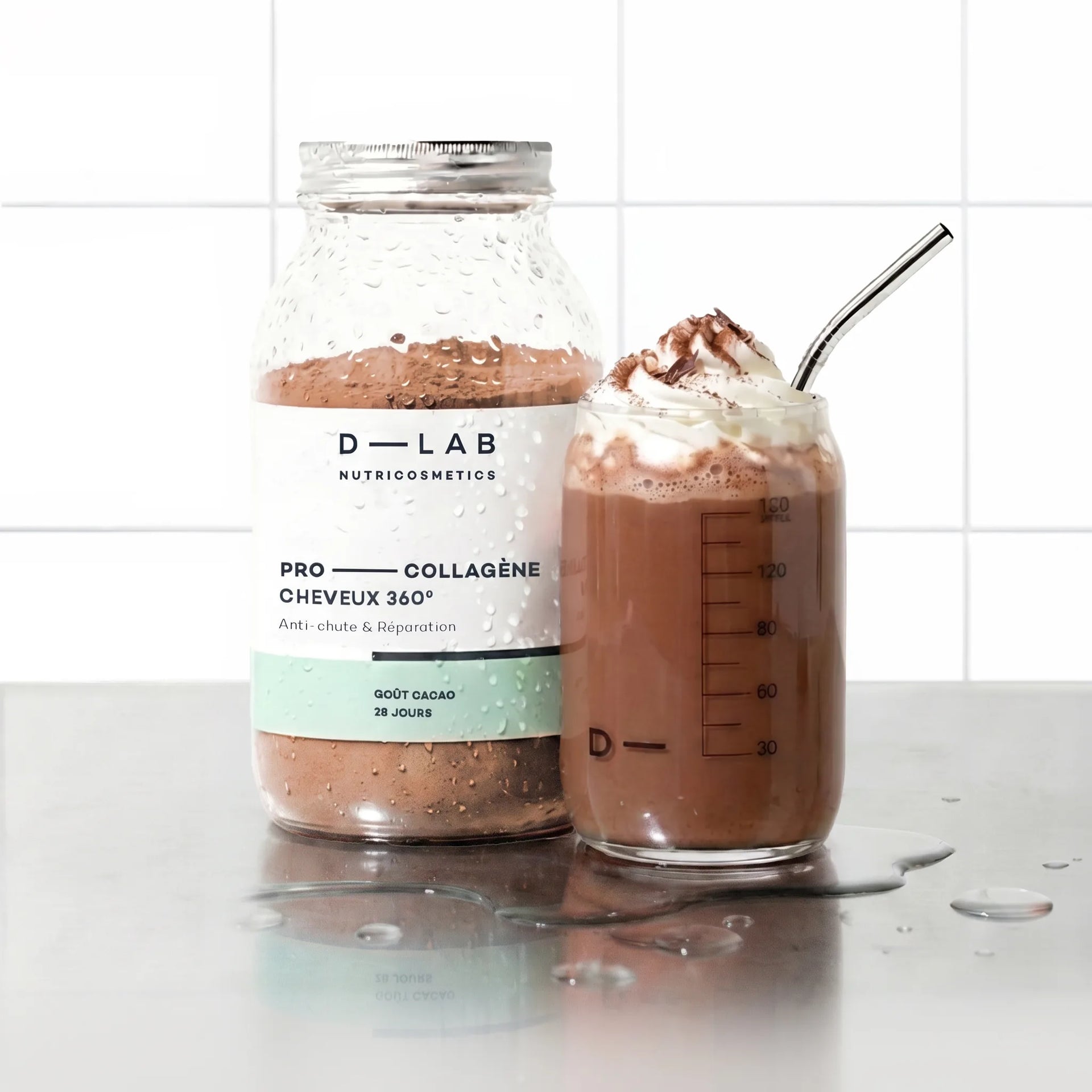 Flacon en verre Pro-Collagène Cheveux 360 goût cacao D-LAB et verre de boisson chocolatée moussée avec paille inox, sur plan de travail carrelé blanc