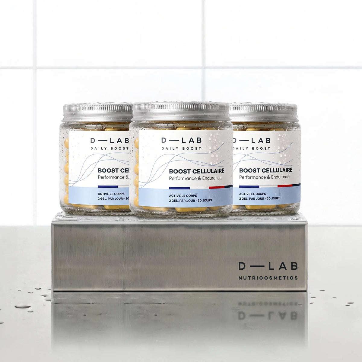 Boost Cellulaire Daily Boost D-LAB Nutricosmetics, cure 3 mois : 3 flacons de gélules alignés sur un socle inox brossé, fond carrelage blanc