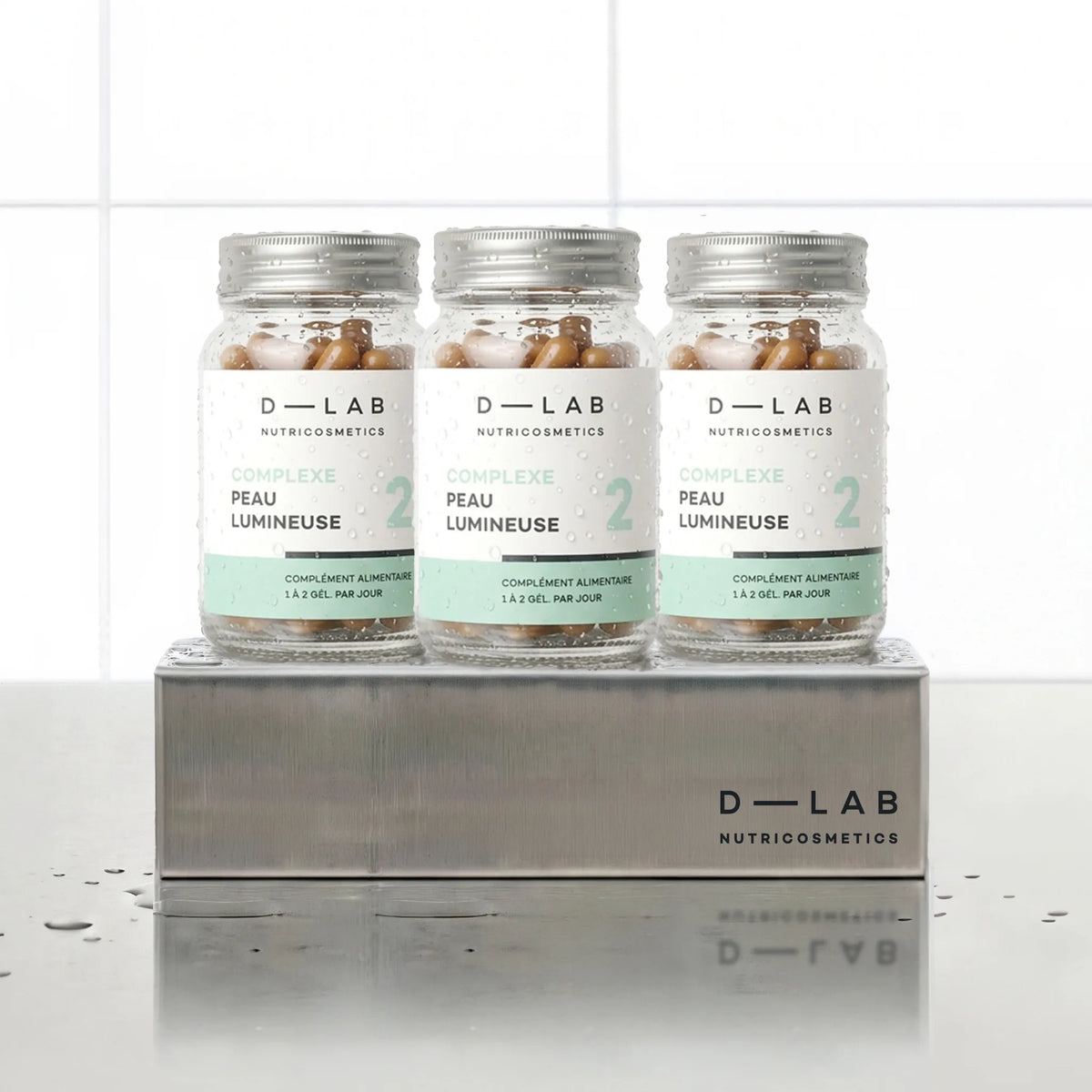 Complexe Peau Lumineuse D-LAB Nutricosmetics, cure 3 mois : 3 flacons de gélules alignés sur un socle inox brossé, fond carrelage blanc
