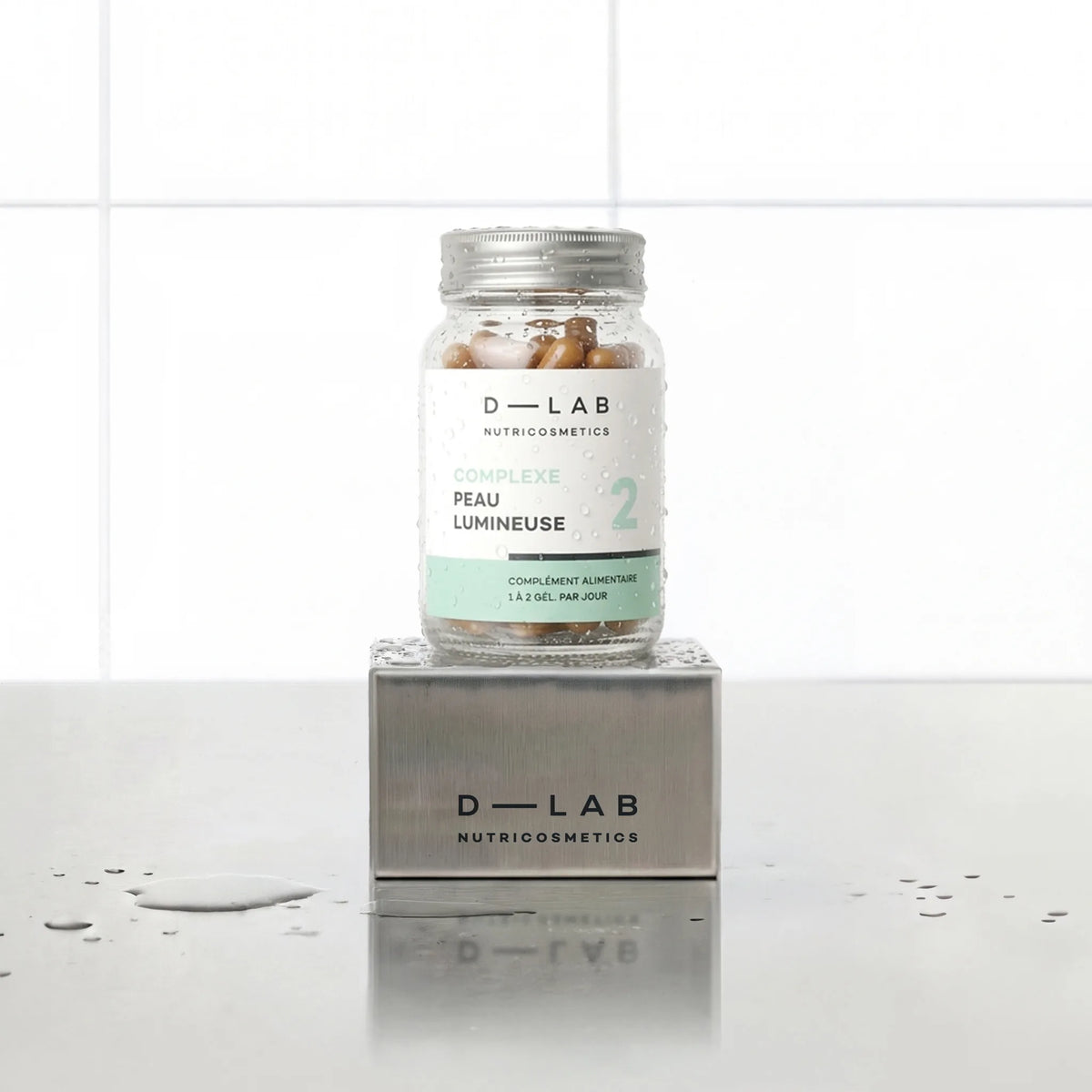 Complexe Peau Lumineuse D-LAB Nutricosmetics, cure 1 mois : flacon de gélules posé sur un socle inox brossé, fond carrelage blanc