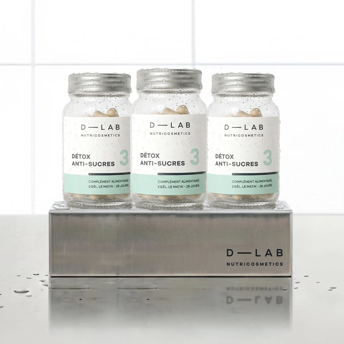 Détox Anti-Sucres D-LAB Nutricosmetics, cure 3 mois : 3 flacons de gélules alignés sur un socle inox brossé, fond carrelage blanc