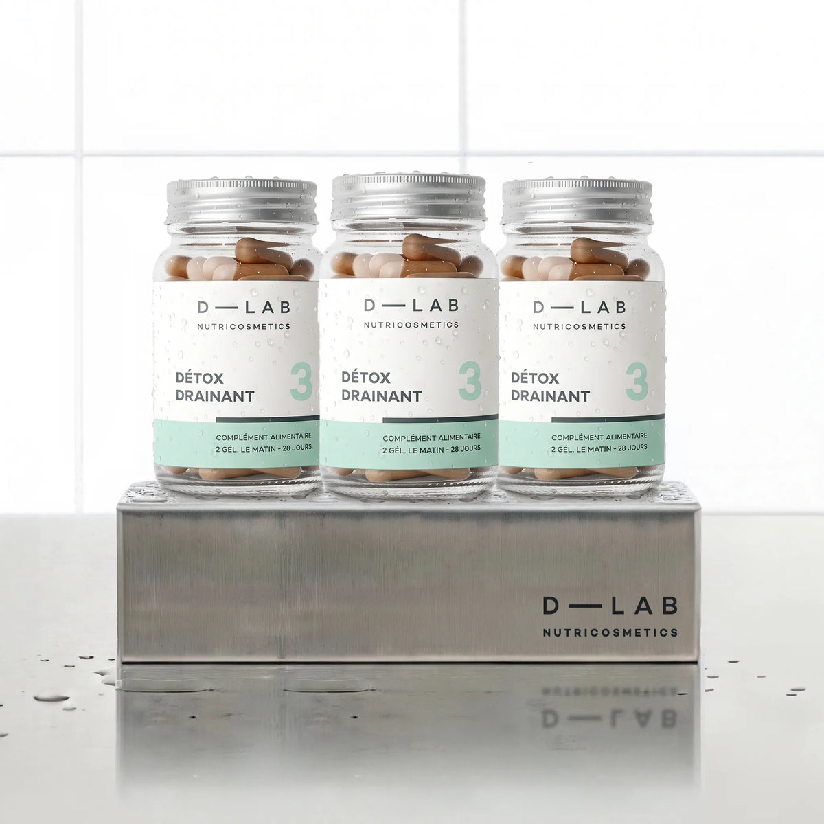 Détox Drainant D-LAB Nutricosmetics, cure 3 mois : 3 flacons de gélules alignés sur un socle inox brossé, fond carrelage blanc
