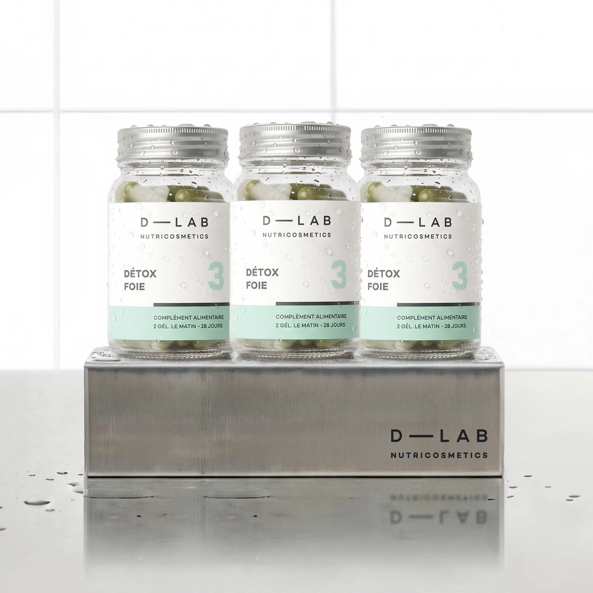 Détox Foie D-LAB Nutricosmetics, cure 3 mois : 3 flacons de gélules alignés sur un socle inox brossé, fond carrelage blanc