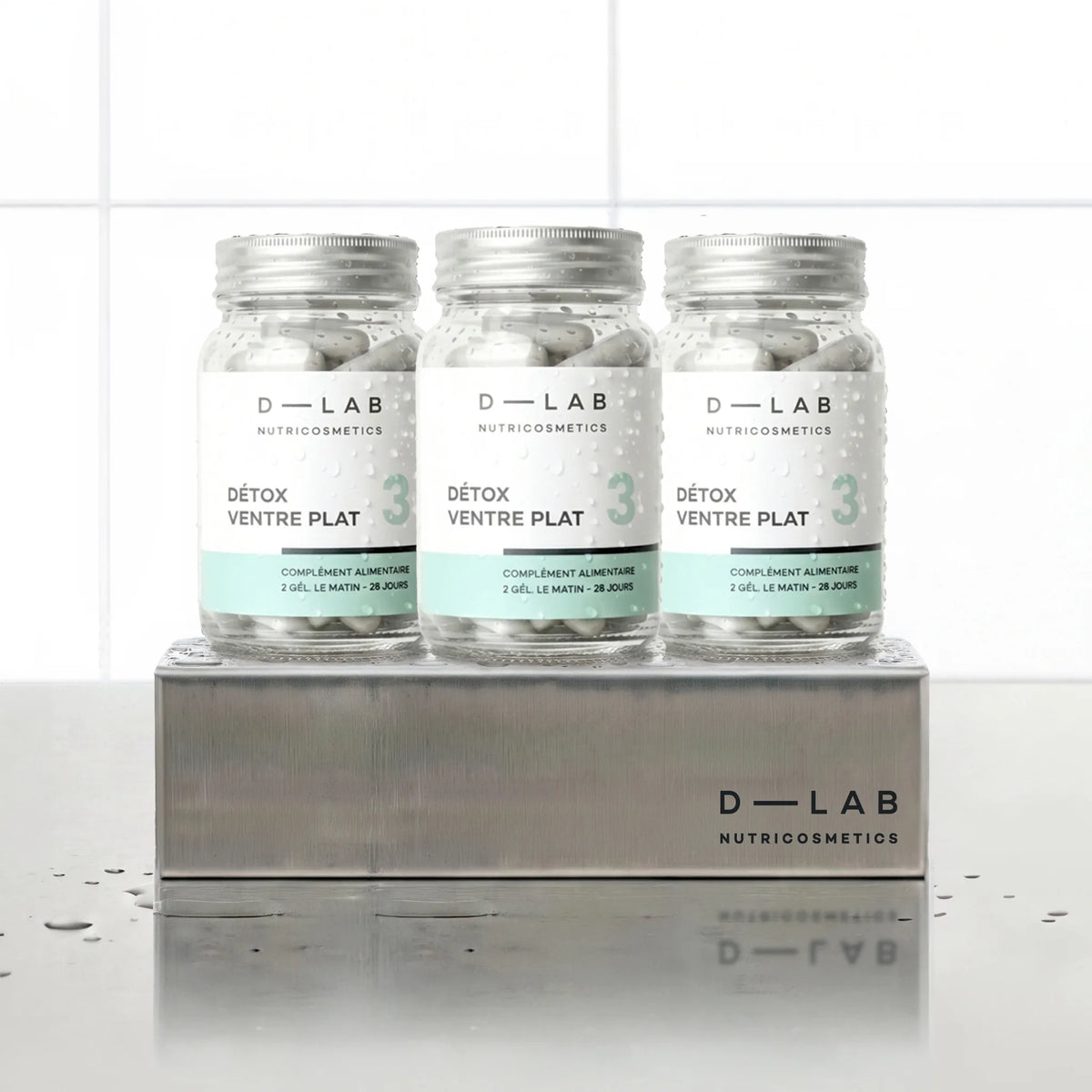 Détox Ventre Plat D-LAB Nutricosmetics, cure 3 mois : 3 flacons de gélules alignés sur un socle inox brossé, fond carrelage blanc